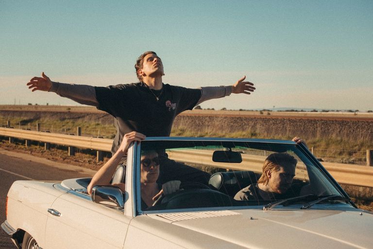 Australian rock band Slowly Slowly members riding in a white convertible car with one member standing up and stretching his arms